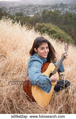 Stock Photography - Portrait of young woman playing acoustic guitar on grassy hill. Fotosearch