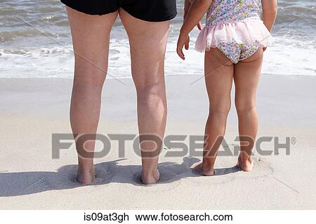 Stock Image - Rear cropped view of senior woman and granddaughters legs on beach. Fotosearch