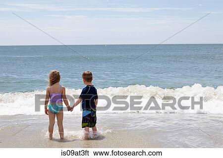 Stock Photograph - Rear view of girl and boy holding hands in sea. Fotosearch