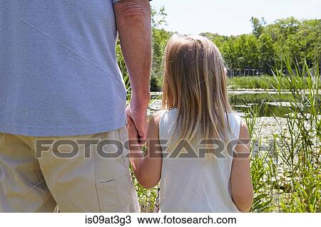 Stock Image - Rear view of girl and grandfather holding hands by rural lake. Fotosearch