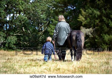 Stock Image - Rear view of mother and son walking pony in field. Fotosearch