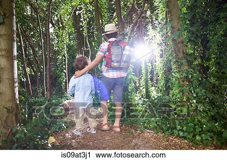 Stock Image - Rear view of two brothers carrying teddy bear and backpack walking in forest. Fotosearch