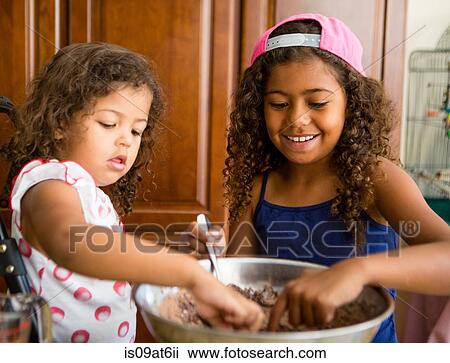 Stock Image - Sisters dipping fingers into chocolate cake mix looking down smiling. Fotosearch