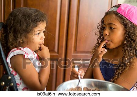 Stock Image - Sisters face to face fingers in mouth tasting chocolate cake mix. Fotosearch