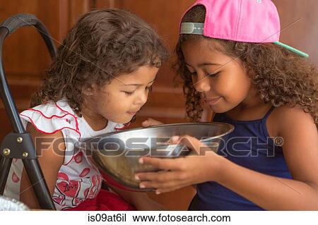 Stock Image - Sisters holding mixing bowl looking down smelling cake mix. Fotosearch