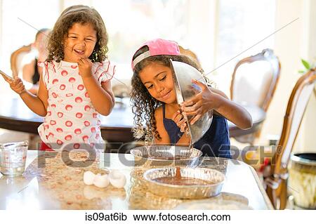 Stock Image - Sisters pouring chocolate cake mix from mixing bowl into cake tins smiling. Fotosearch