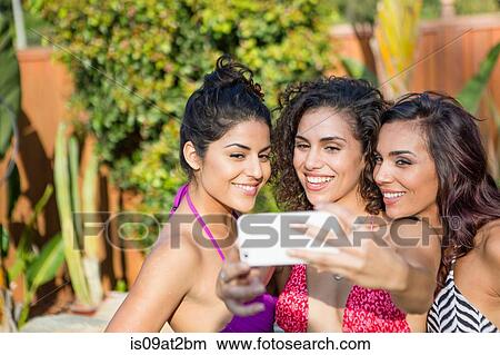 Picture - Three adult sisters wearing bikini tops posing for smartphone selfie in garden. Fotosearch