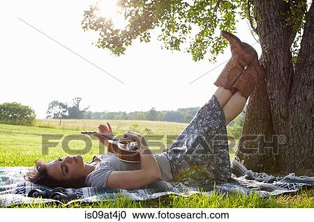 Stock Image - Woman lying on picnic blanket in field playing ukuele. Fotosearch