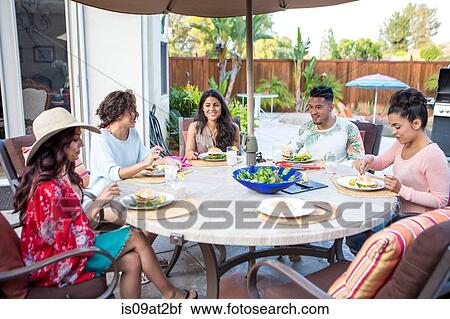 Stock Image - Young man and four adult sisters having family barbecue lunch on patio. Fotosearch
