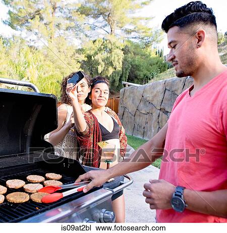 Young man cooking on barbecue in front of sisters taking smartphone selfie View Large Photo Image Stock Image - Young man cooking on barbecue in front of sisters taking smartphone selfie. Fotosearch