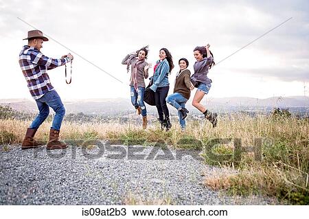 Stock Image - Young man photographing his four adult sisters jumping on rural hill. Fotosearch