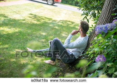 Stock Image - "Young man sitting against tree, using laptop". Fotosearch