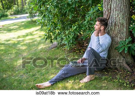 Stock Image - "Young man sitting against tree, using laptop". Fotosearch