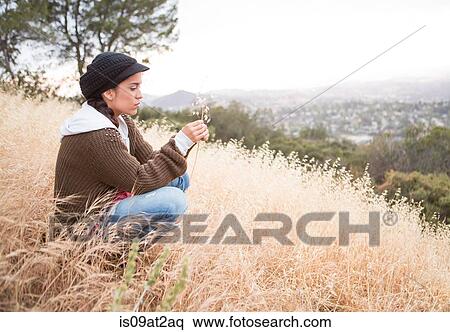 Young woman crouching on grassy hill holding dry blade of grass View Large Photo Image Stock Photography - Young woman crouching on grassy hill holding dry blade of grass. Fotosearch