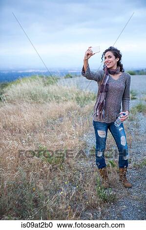 Stock Image - Young woman posing for smartphone selfie on rural hill. Fotosearch