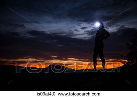 Stock Image - Young woman standing at nights in darkness wearing headlamp. Fotosearch