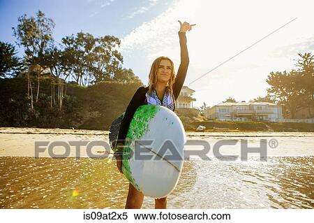 Stock Photography - "Young woman standing in sea, looking out to sea, holding surfboard, gesturing with hand". Fotosearch
