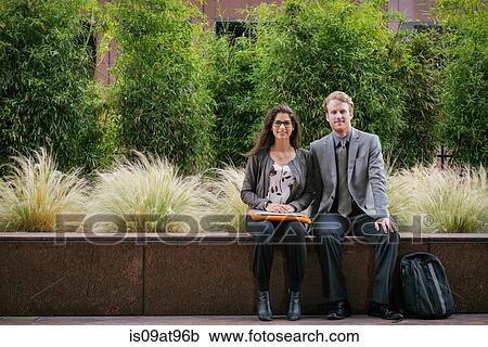 Stock Image - Businessman and woman in city sitting on wall with laptop. Fotosearch
