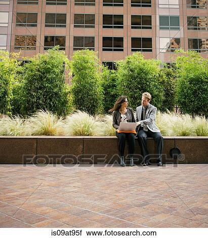 Stock Image - Businessman and woman sitting on wall using laptop. Fotosearch