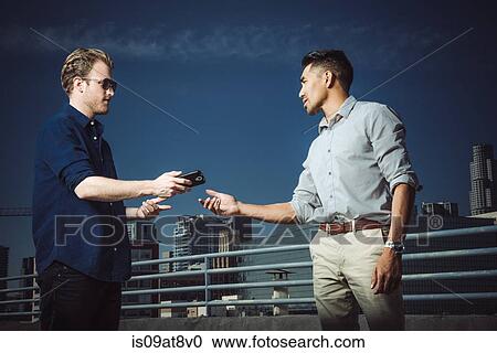 Stock Image - Businessman giving smartphone to man on roof terrace, Los Angeles, California, USA. Fotosearch