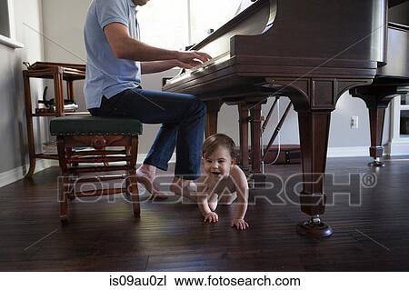 Stock Image - Father playing piano with baby boy crawling at feet. Fotosearch