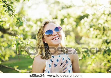 Stock Image - Head and shoulders of young woman wearing sunglasses, hand on chest looking at camera laughing. Fotosearch