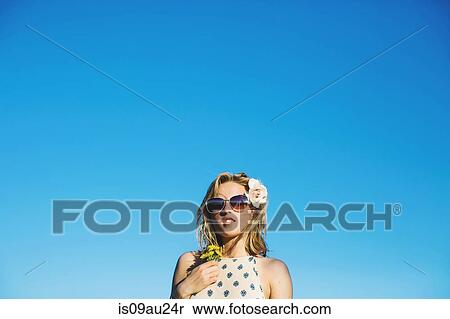 Stock Photography - Low angle view of young woman weraing sunglasses, flowers in hair holding dandelion against blue sky. Fotosearch
