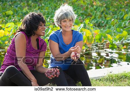 Stock Image - Mature female friends sitting in park, eating grapes. Fotosearch