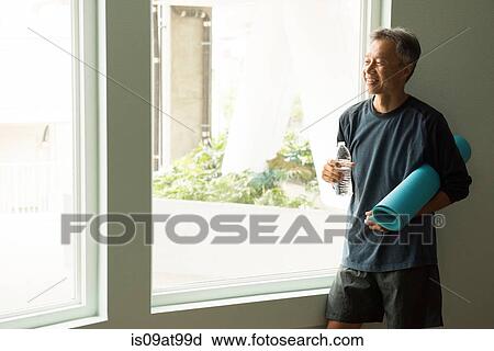 Stock Image - Mature man holding rolled up yoga mat and bottle of water looking out of window. Fotosearch