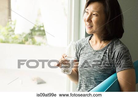 Mature woman holding rolled up yoga mat and bottle of water looking out of window smiling View Large Photo Image Stock Image - Mature woman holding rolled up yoga mat and bottle of water looking out of window smiling. Fotosearch