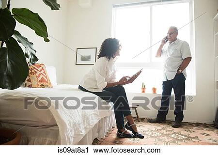 Stock Image - Mature woman on bed using digital tablet whilst husband talks on smartphone. Fotosearch