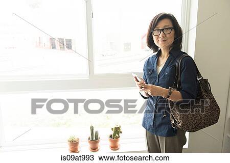 Mature woman standing in front of window holding smartphone looking at camera smiling View Large Photo Image Stock Image - Mature woman standing in front of window holding smartphone looking at camera smiling. Fotosearch