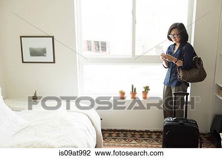 Mature woman standing in hotel room with suitcase using smartphone View Large Photo Image Stock Image - Mature woman standing in hotel room with suitcase using smartphone. Fotosearch