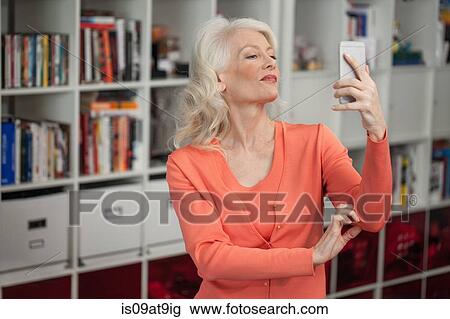 Stock Image - Mature woman taking self portrait, using smartphone, indoors. Fotosearch