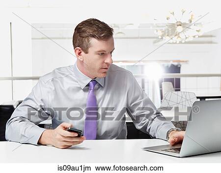 Picture - Mid adult businessman sitting at desk, using laptop. Fotosearch