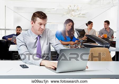 Mid adult businessman sitting at desk, using laptop, colleagues working in background View Large Photo Image Picture - Mid adult businessman sitting at desk, using laptop, colleagues working in background. Fotosearch