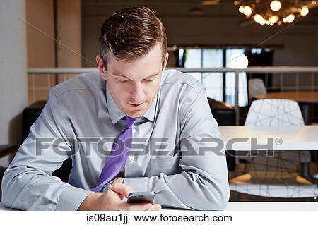 Mid adult businessman sitting at desk, looking at smartphone View Large Photo Image Stock Image - Mid adult businessman sitting at desk, looking at smartphone. Fotosearch