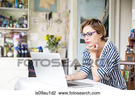 Stock Image - Mid adult woman working on laptop at kitchen table. Fotosearch