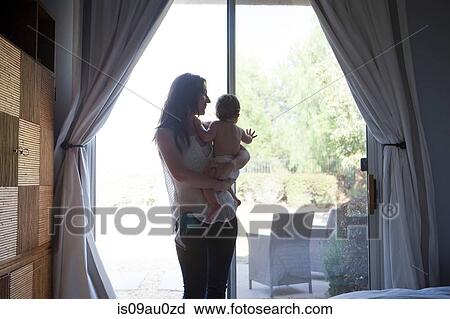 Stock Image - Mother holding baby boy in front of patio door, looking away. Fotosearch