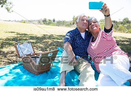 Senior couple sitting on picnic blanket, taking self portrait using smartphone View Large Photo Image Stock Photo - Senior couple sitting on picnic blanket, taking self portrait using smartphone. Fotosearch