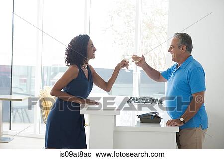 Stock Image - Senior man and wife raising a glass of white wine over kitchen counter. Fotosearch