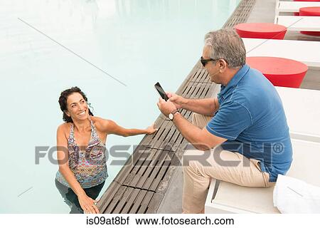 Stock Image - Senior man photographing wife with smartphone from poolside. Fotosearch