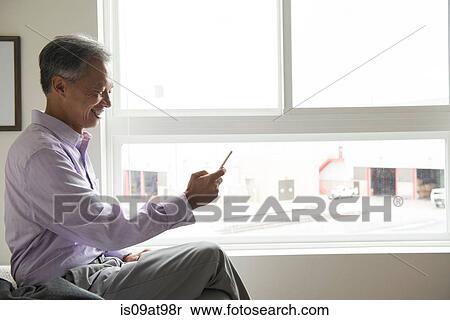 Side view of mature man sitting in front of window using smartphone View Large Photo Image Stock Photography - Side view of mature man sitting in front of window using smartphone. Fotosearch