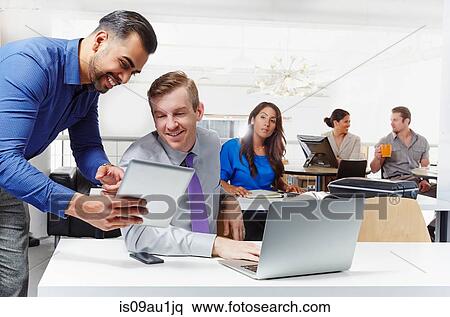 Stock Photography - Two businessmen having discussion, looking at digital tablet, colleagues working in background. Fotosearch
