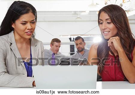 Two businesswomen sitting at desk, looking at laptop View Large Photo Image Stock Photo - Two businesswomen sitting at desk, looking at laptop. Fotosearch