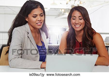 Two businesswomen sitting at desk, looking at laptop View Large Photo Image Stock Photo - Two businesswomen sitting at desk, looking at laptop. Fotosearch