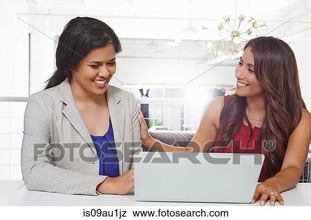 Two businesswomen sitting at desk, looking at laptop View Large Photo Image Stock Photo - Two businesswomen sitting at desk, looking at laptop. Fotosearch