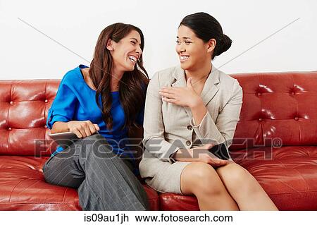 Stock Image - Two businesswomen sitting on sofa, having discussion, laughing. Fotosearch