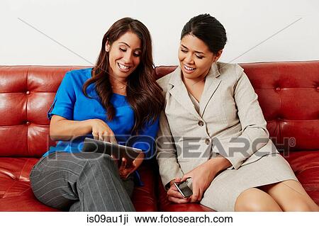 Two businesswomen sitting on sofa, looking at digital tablet View Large Photo Image Stock Image - Two businesswomen sitting on sofa, looking at digital tablet. Fotosearch
