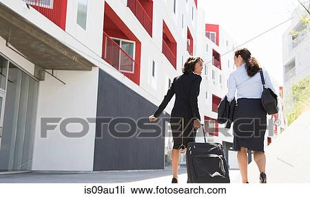 Two businesswomen walking along street, pulling suitcase, rear view View Large Photo Image Stock Image - Two businesswomen walking along street, pulling suitcase, rear view. Fotosearch